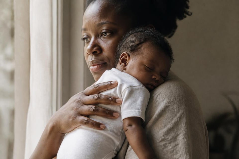 A woman holds a sleeping baby at a window.